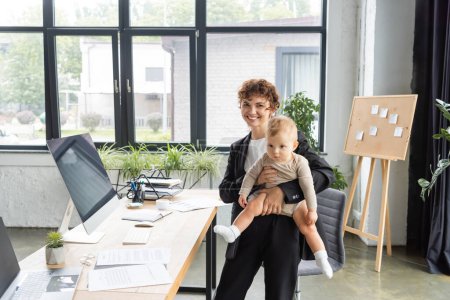Photo for Happy businesswoman standing with baby girl near work desk with computers and documents in modern office - Royalty Free Image