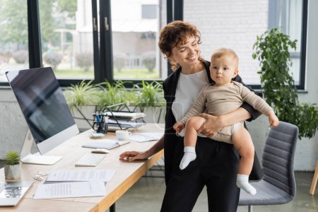 smiling businesswoman holding toddler daughter in romper near work desk with computer monitor and documents