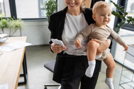 smiling businesswoman holding little daughter and using mobile phone in office