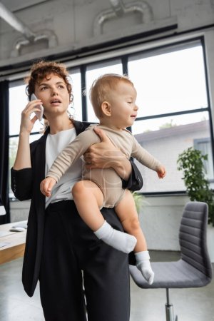 businesswoman in black suit talking on cellphone while holding toddler daughter in office