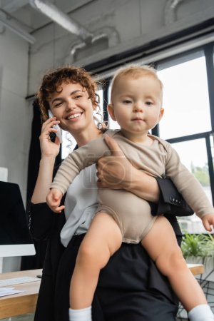 positive businesswoman talking on smartphone and holding toddler child in office