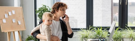 businesswoman talking on mobile phone and holding little daughter near computer monitor in office, banner
