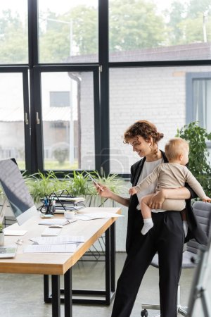Photo for Happy businesswoman standing with smartphone and baby near work desk with documents and computer monitor with blank screen - Royalty Free Image