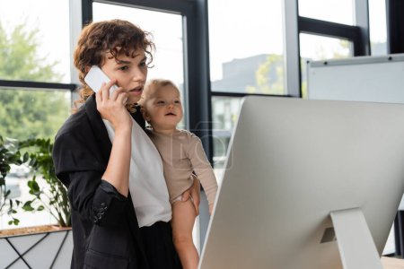 Photo for Woman in black suit holding smiling baby while talking on cellphone near computer monitor - Royalty Free Image