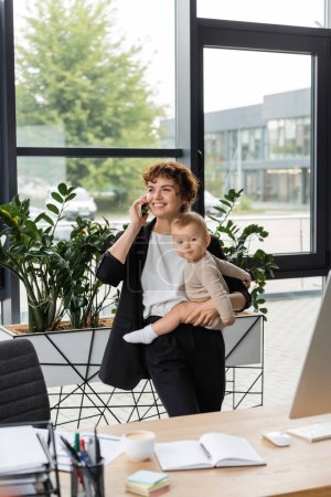 Photo for Happy businesswoman looking away while holding baby and talking on mobile phone in modern office - Royalty Free Image
