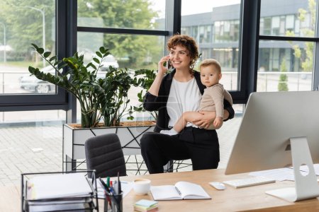 happy businesswoman holding toddler child and talking on smartphone near work desk and green plants in modern office