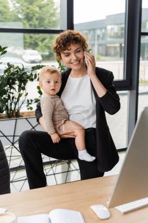 joyful businesswoman holding toddler daughter and talking on smartphone near computer monitor in office
