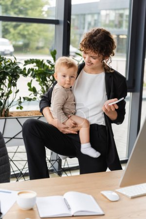 Téléchargez les photos : Smiling businesswoman with baby and mobile phone near blurred notebook and coffee cup on work desk - en image libre de droit