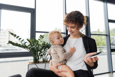 Photo for Pleased businesswoman holding baby and mobile phone with blank screen near green plants in office - Royalty Free Image