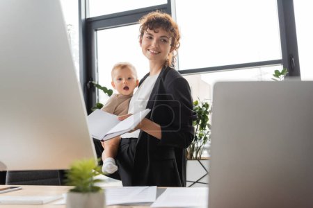Photo for Smiling businesswoman holding baby and blank notebook near blurred computers in office - Royalty Free Image