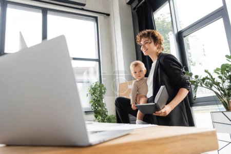 happy businesswoman in black suit holding toddler child and notebook near laptop on blurred foreground