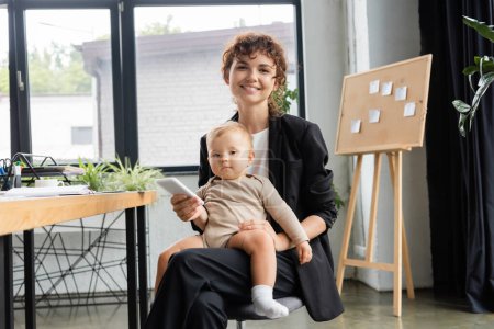 happy businesswoman sitting with little daughter and mobile phone while looking at camera in office