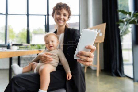 Photo for Blurred businesswoman taking selfie on mobile phone while sitting with smiling baby in office - Royalty Free Image