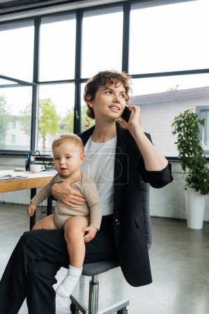 Photo for Positive businesswoman looking away while sitting with baby in office and talking on smartphone - Royalty Free Image