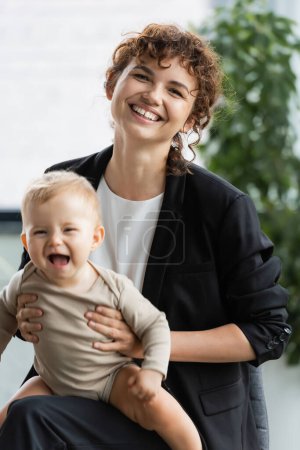 businesswoman in black suit smiling at camera while holding excited baby in office
