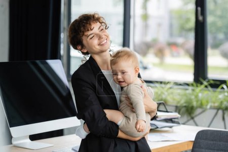 cheerful businesswoman in black blazer holding toddler child near computer monitor with blank screen in office