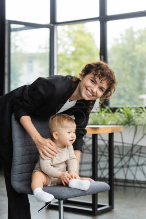 cheerful businesswoman smiling at camera near toddler child sitting on office chair
