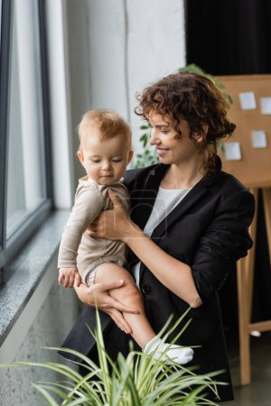 smiling businesswoman in black blazer standing with toddler child near blurred plant in office