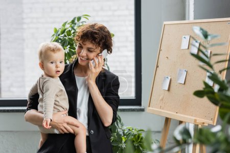 happy businesswoman talking on mobile phone while standing with baby near cork board with paper notes