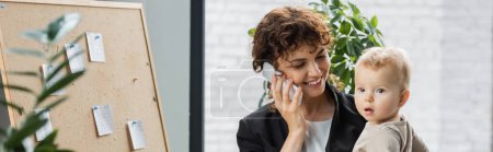 happy businesswoman calling on mobile phone near little child and cork board with paper notes in office, banner