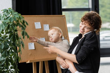 smiling businesswoman in black blazer talking on smartphone near little daughter touching leaves of green plant in office