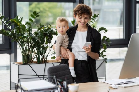 smiling businesswoman holding toddler daughter and using mobile phone at workplace in office