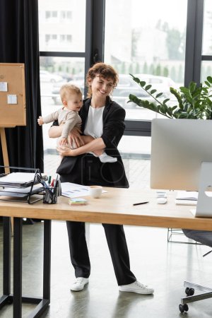 happy businesswoman in black suit looking at computer monitor while standing with little child in office