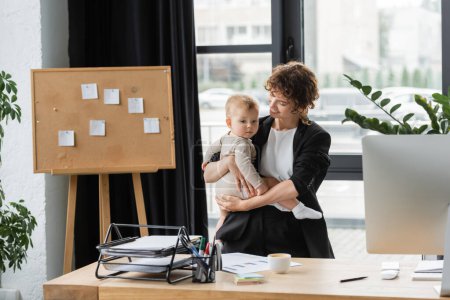 smiling businesswoman holding toddler daughter near work desk and cork board with paper notes in office