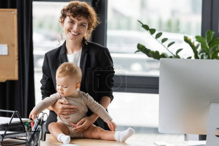businesswoman in black blazer smiling at camera near little daughter in romper sitting on office desk