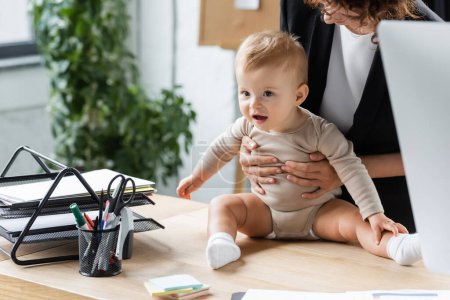 businesswoman holding excited baby sitting on office desk near documents and stationery