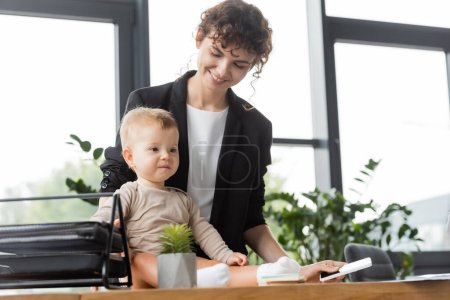 happy businesswoman in black blazer holding mobile phone near toddler daughter sitting on office desk