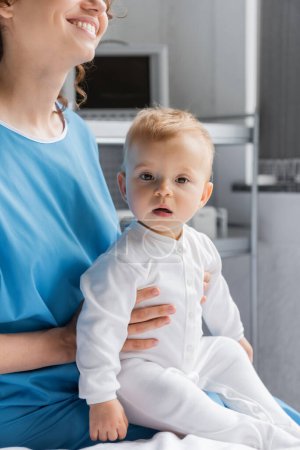cheerful woman in patient gown holding little daughter looking at camera in clinic