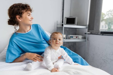 toddler child in romper sitting near mom in patient gown looking away on bed in clinic