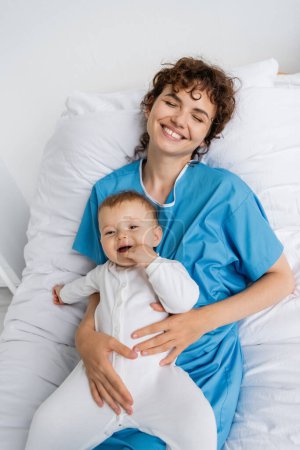 top view of happy woman holding little daughter and smiling with closed eyes on hospital bed