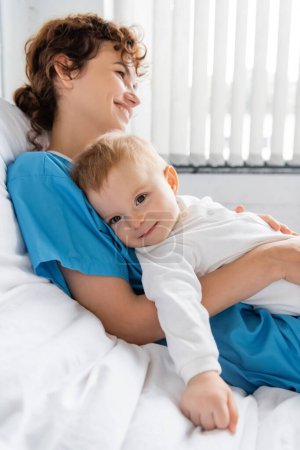 happy woman holding smiling child in romper on bed in hospital ward