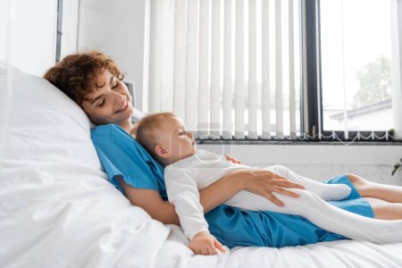 pleased woman in patient gown lying on hospital bed with toddler child in romper