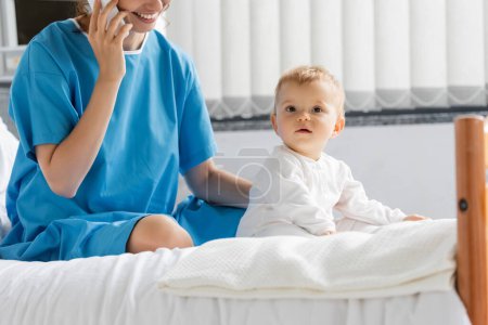 toddler girl in romper sitting on hospital bed near smiling mom in patient gown talking on mobile phone