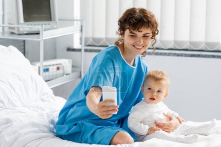 cheerful woman in patient gown taking selfie with little daughter on bed in hospital ward