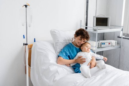 joyful woman in patient gown hugging little baby in romper while sitting on hospital bed