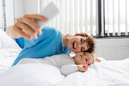 excited woman with mobile phone taking selfie with toddler child on bed in hospital ward