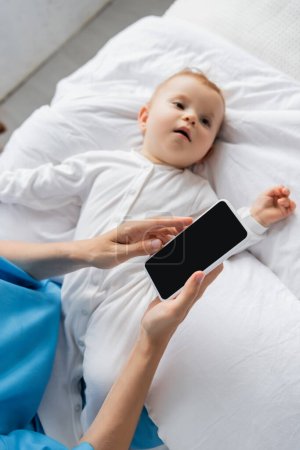 top view of blurred baby in romper lying on hospital bed near mom holding cellphone with blank screen