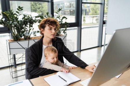 businesswoman in black blazer sitting at workplace with little daughter and working on computer near blank notebook