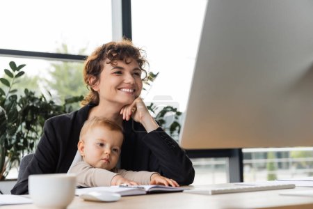 smiling businesswoman looking at computer monitor while working in office near toddler daughter