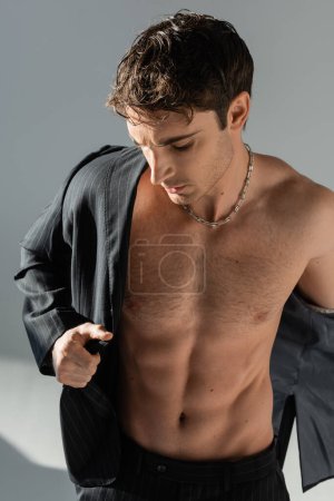 shirtless man in silver necklace and black jacket undressing on grey background