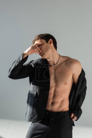 Téléchargez les photos : Shirtless man in black blazer and pants covering face with hand while standing on grey background - en image libre de droit