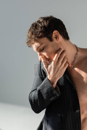 Photo for Sexy man in silver necklace and blazer on shirtless torso touching face on grey background - Royalty Free Image