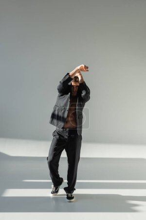Foto de Full length of trendy man in black suit and sneakers covering head with blazer while posing with raised hands on grey background - Imagen libre de derechos