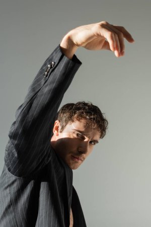 Foto de Brunette man in black and striped blazer posing with raised hand while looking at camera isolated on grey - Imagen libre de derechos