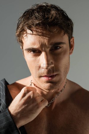 Foto de Portrait of sexy brunette man touching silver necklace and looking at camera isolated on grey - Imagen libre de derechos
