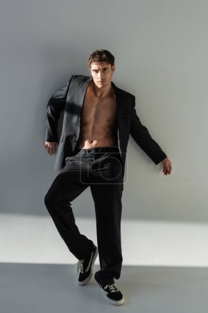 full length of sexy shirtless man in black suit and sneakers standing on grey background with lighting